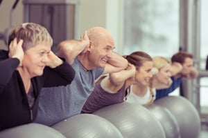 Happy elderly couple exercising in a pilates class at the gym with three other younger people toning and strengthening their muscles using gym balls, focus to the senior man and woman